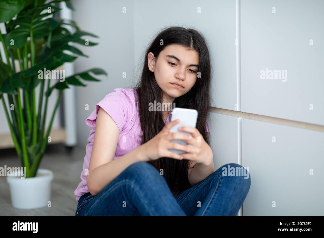 Deprimiertes indisches Teenager-Mädchen mit Smartphone, das sich einsam fühlt, auf den Bildschirm schaut und zu Hause in der Nähe der Wand sitzt Stockfoto