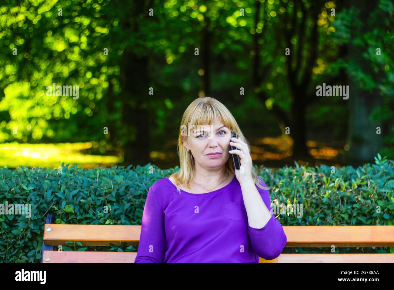 Unschärfe kaukasisch blonde Frau reden, sprechen am Telefon draußen, im Freien. 40-jährige Frau in lila Bluse im Park auf der Bank. Erwachsene weibliche Erbsenw Stockfoto