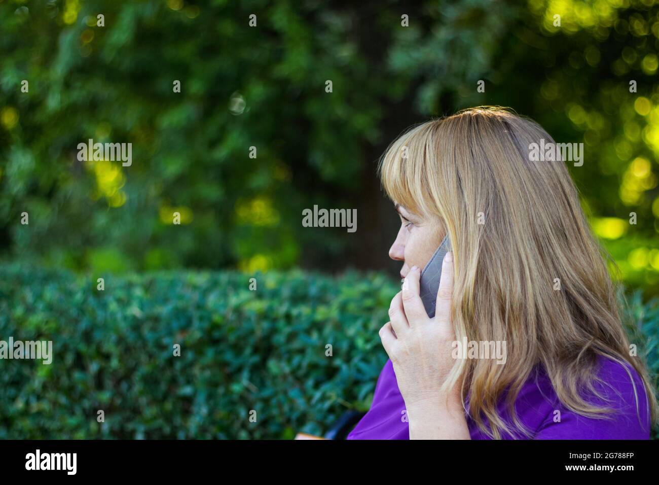 Unschärfe kaukasisch blonde Frau sprechen auf dem Telefon draußen, draußen. 40er Jahre alte Frau in lila Bluse im Park, Profil. Seriöse Person, die mobil verwendet Stockfoto