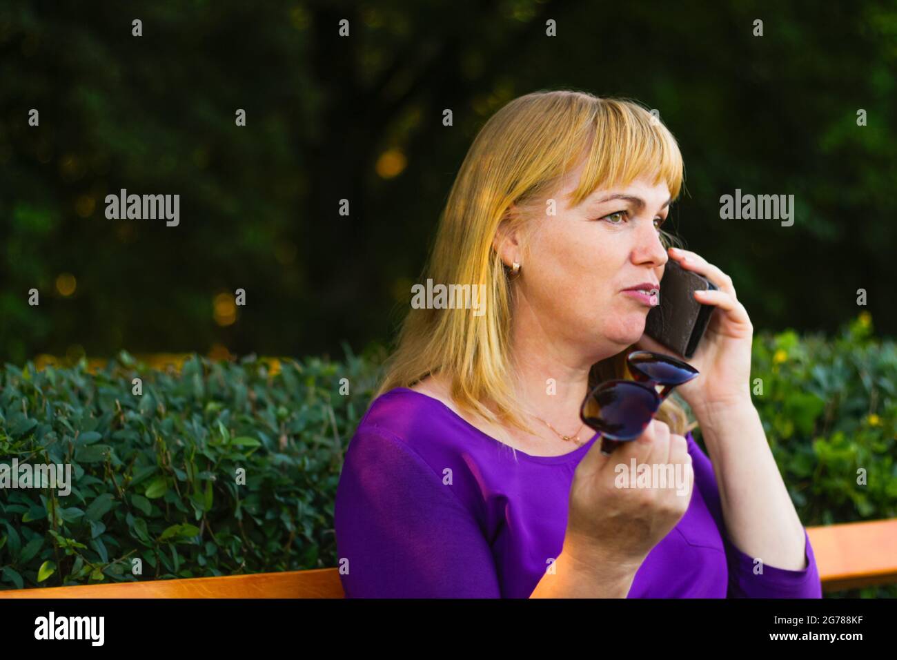 Unschärfe-Nahaufnahme kaukasisch blonde Frau sprechen, sprechen auf dem Telefon draußen, im Freien. 40-jährige Frau in lila Bluse im Park. Erwachsene Menschen usi Stockfoto