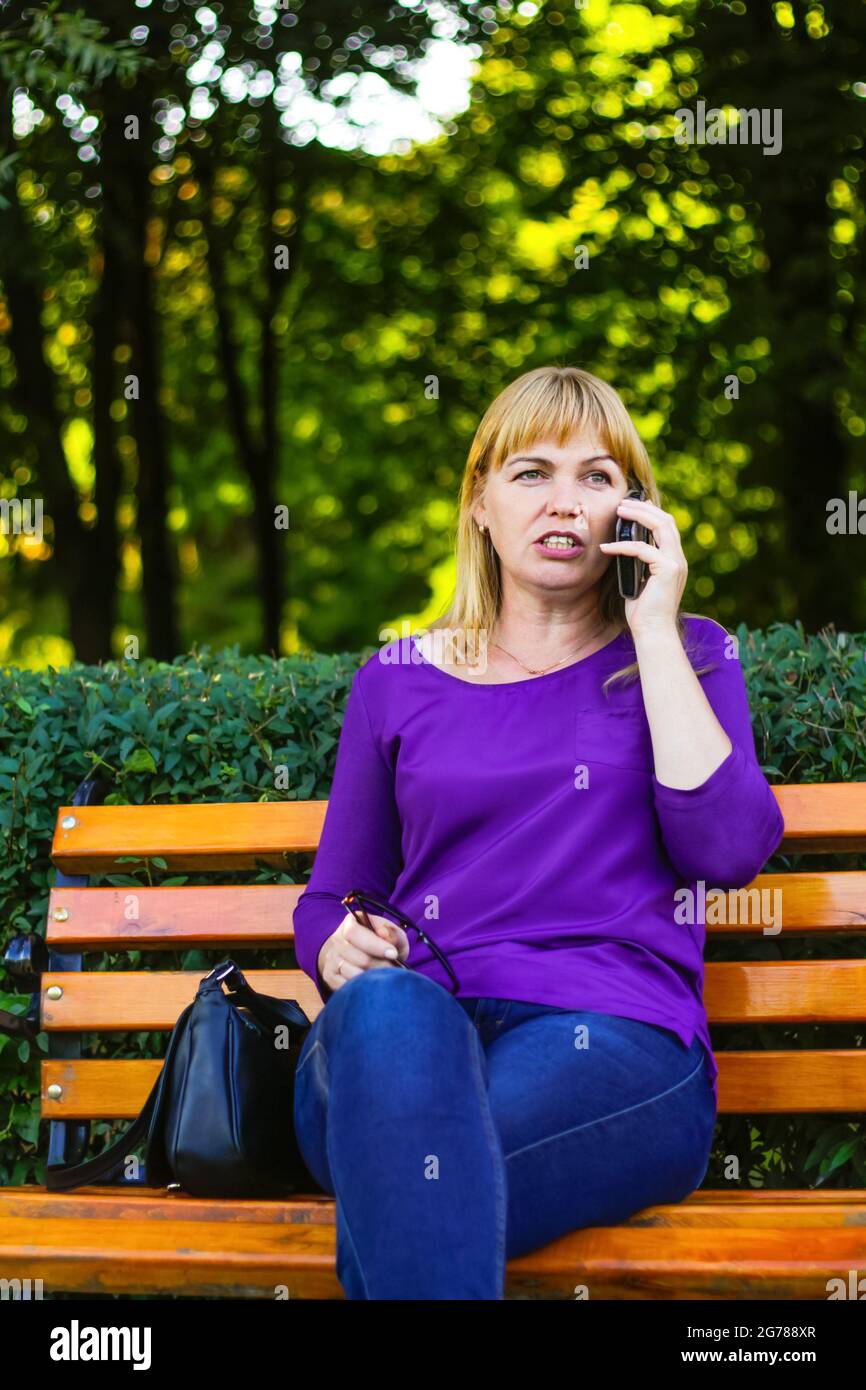 Unschärfe Wunder kaukasische blonde Frau reden, sprechen am Telefon draußen, im Freien. 40-jährige Frau in lila Bluse im Park. Erwachsene Person mit Stockfoto