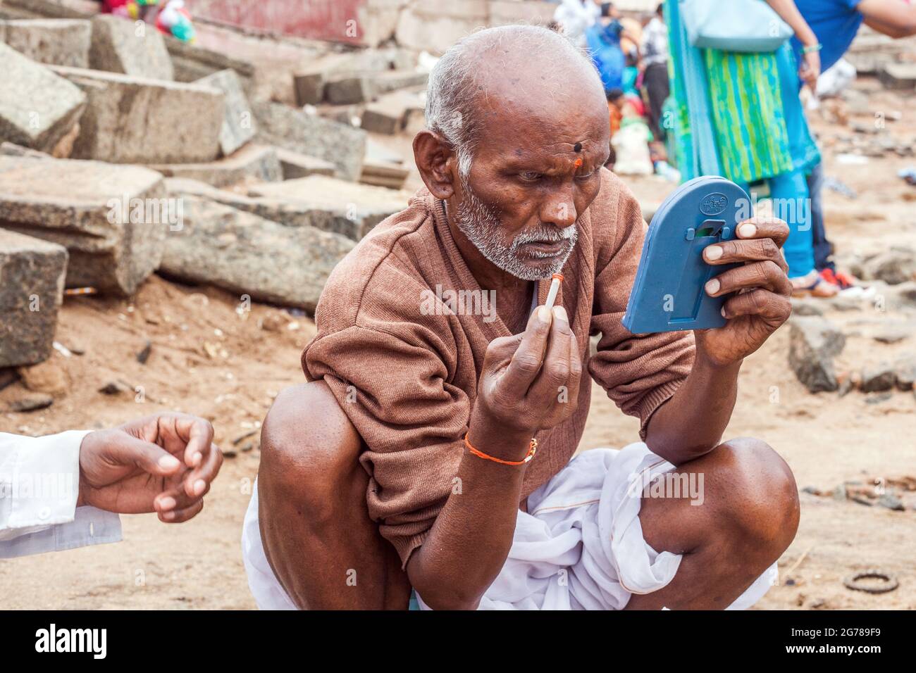 Älterer, kahler indischer Mann, der auf dem Boden hockte und Lunghi trug, trägt Bindi auf seine Stirn, während er in einen Spiegel blickt Stockfoto