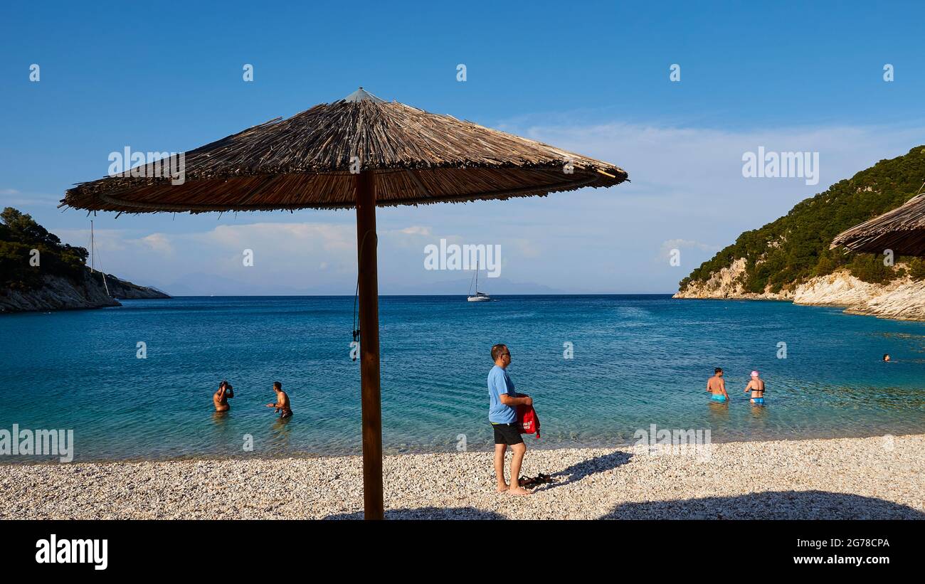 Ionische Inseln, Ithaca, Insel Odysseus, in der Nähe von Vathi, Filiatro Beach, Feiner Kiesstrand, Menschen am Strand, runder Sonnenschirm mit Bambusstroh bedeckt, Menschen im Wasser, blauer Himmel mit Wolken, ein einziges Segelboot ist in der Bucht verankert Stockfoto