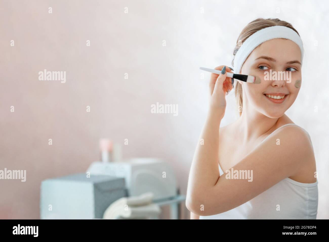 Junge Frau tragen Clay Gesichtsmaske. Teenager-Mädchen breitet Tonmaske auf Wange mit Pinsel zu Hause im Badezimmer. Self Care Beauty Skincare Spa und Behandlung Stockfoto