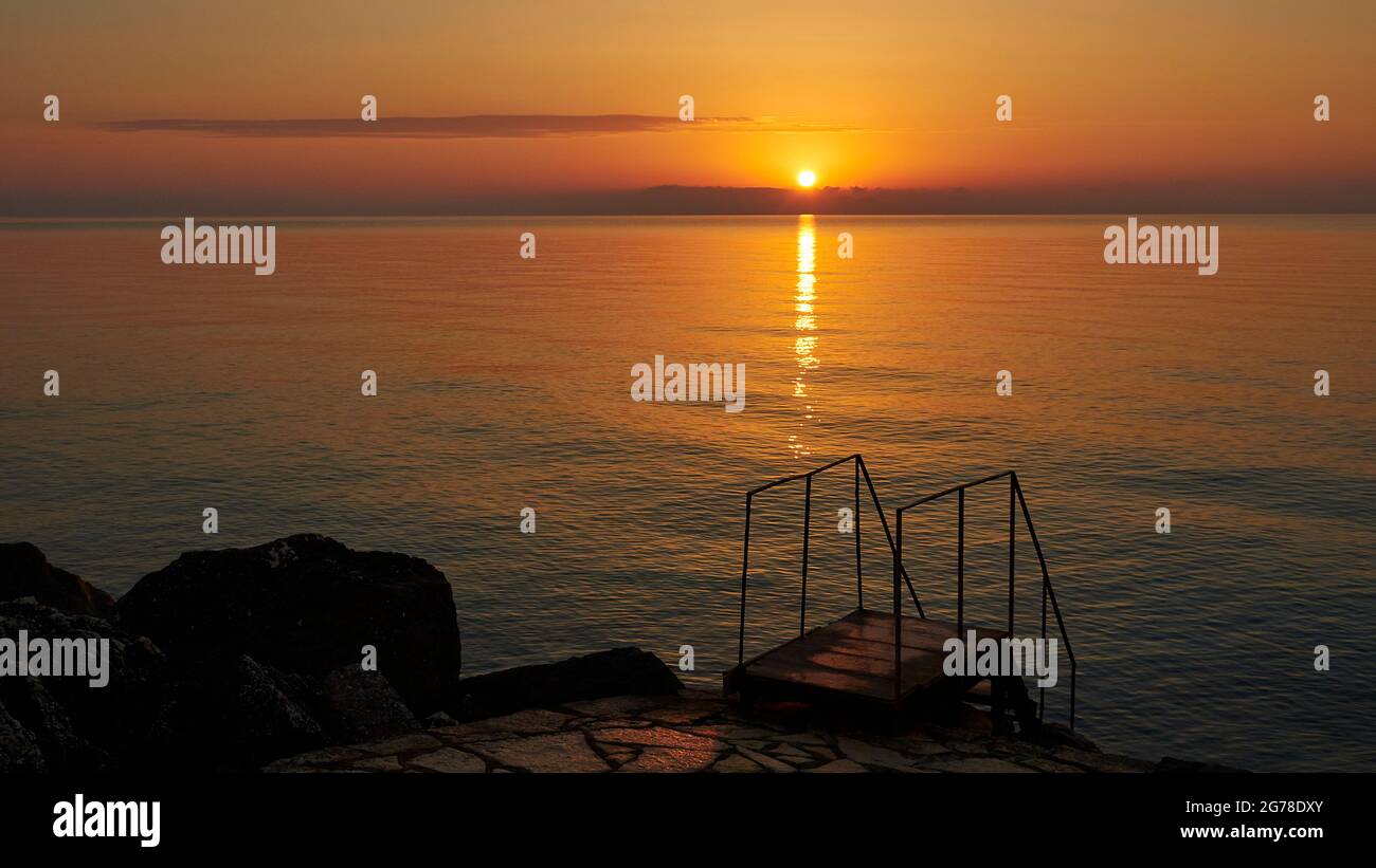 Zakynthos, Zakynthos Stadt, Morgenlicht, Morgenstimmung, Sonnenaufgang über dem Meer, kleine Sonnenscheibe. Im unteren dritten Schatten führen Treppen mit Handläufen ins Wasser Stockfoto