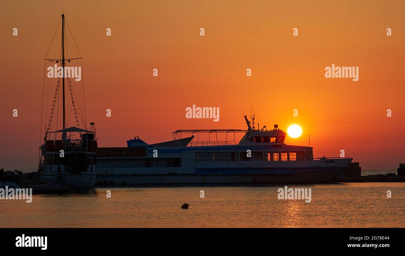 Zakynthos, Zakynthos Stadt, Hafen, Sonnenaufgang, Segelboot links, Ausflugsboot rechts, beides als Silhouette, die Sonne geht hinter dem Ausflugsboot auf. Orangefarbener Morgenhimmel, einzelne Boje im Vordergrund Stockfoto