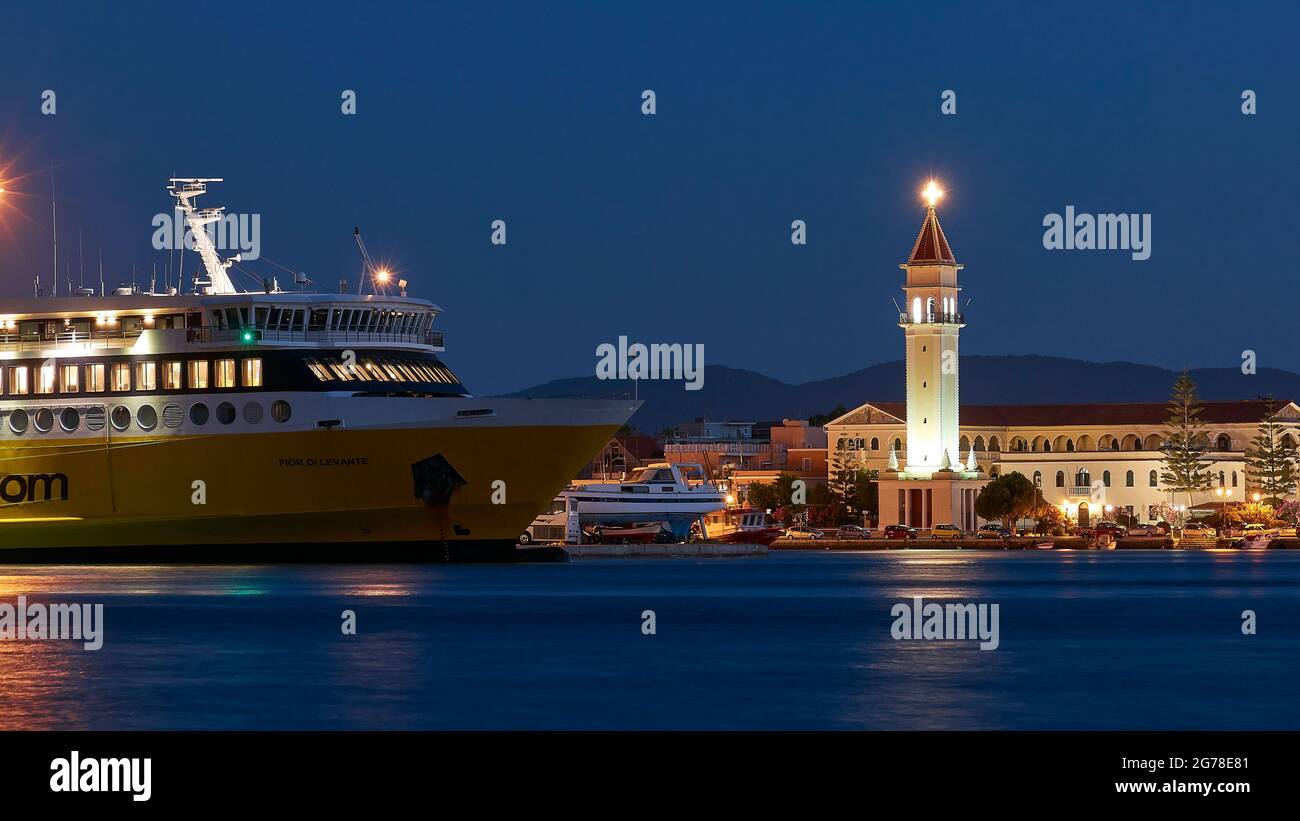 Zakynthos, Zakynthos Stadt, Nachtaufnahme, Abendaufnahme, Hafen, Vorderteil der Zante-Fähre in der mittleren Entfernung, im Vordergrund blaues Wasser, beleuchtete Agios Dionysios-Kirche direkt hinter der Fähre, blauer Nachthimmel, festlich beleuchteter Kirchturm Stockfoto