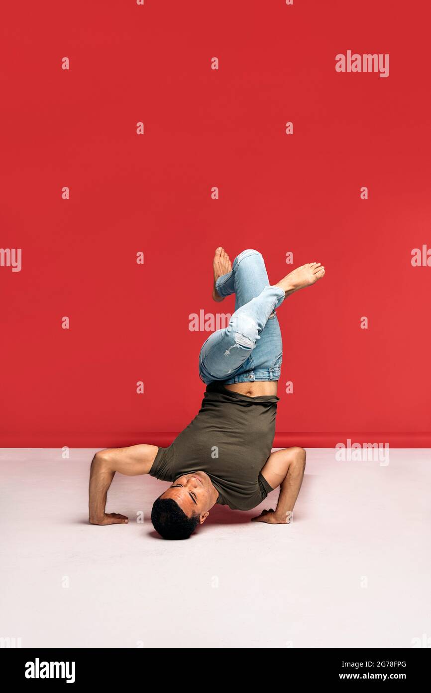 Hispanische Tänzerin mit legerer Kleidung, die Kopfstand macht und im Studio vor rotem Hintergrund tanzt. Stockfoto