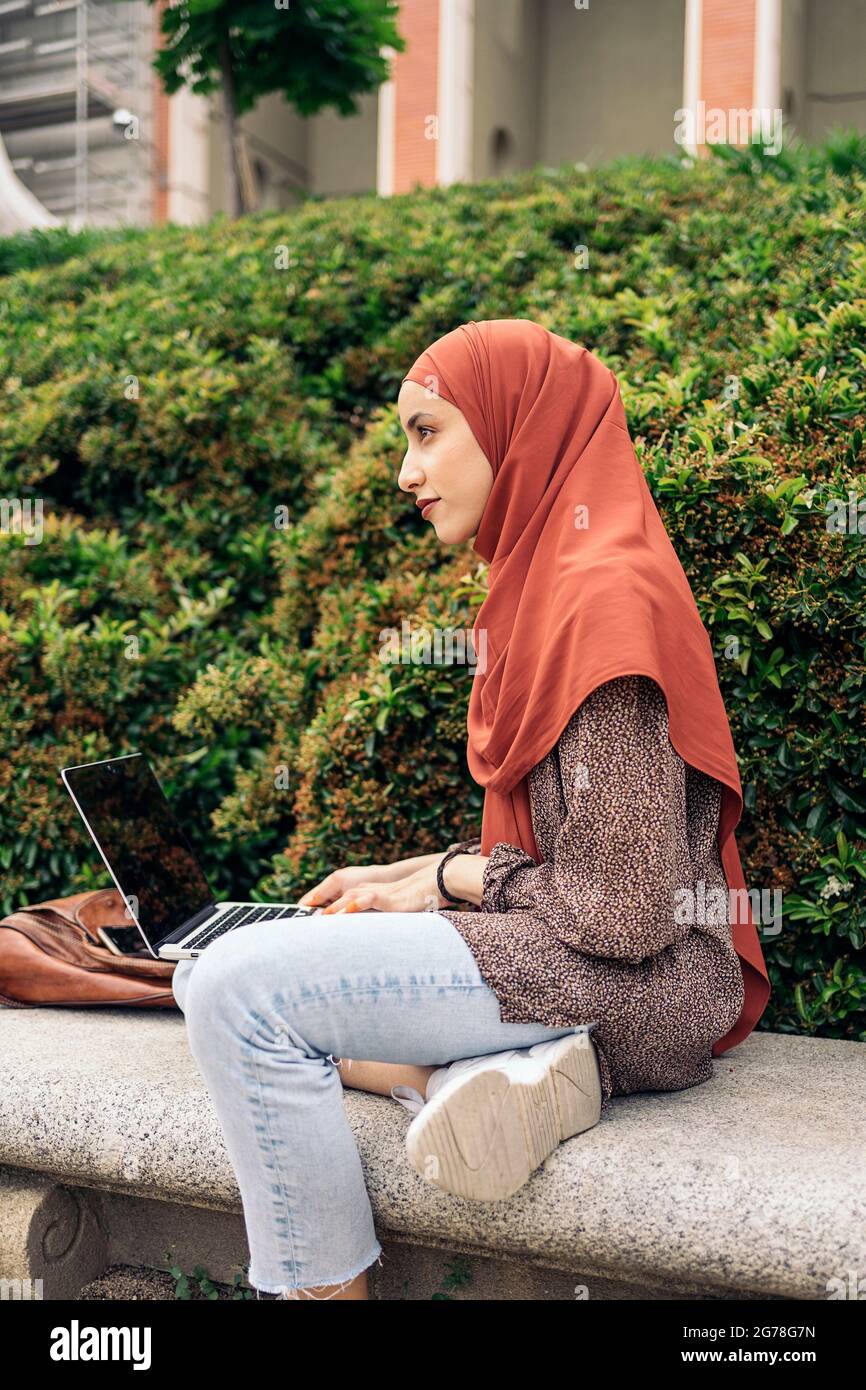 Schöne junge muslimische Frau mit Hijab, die im Park sitzt und ihren Computer benutzt. Stockfoto