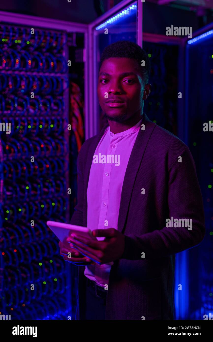 Porträt eines ernsthaften jungen schwarzen Netzwerksicherheitstechnikers in Strickjacke, der mit einem Tablet gegen den Rack-Schrank eines Servers steht Stockfoto