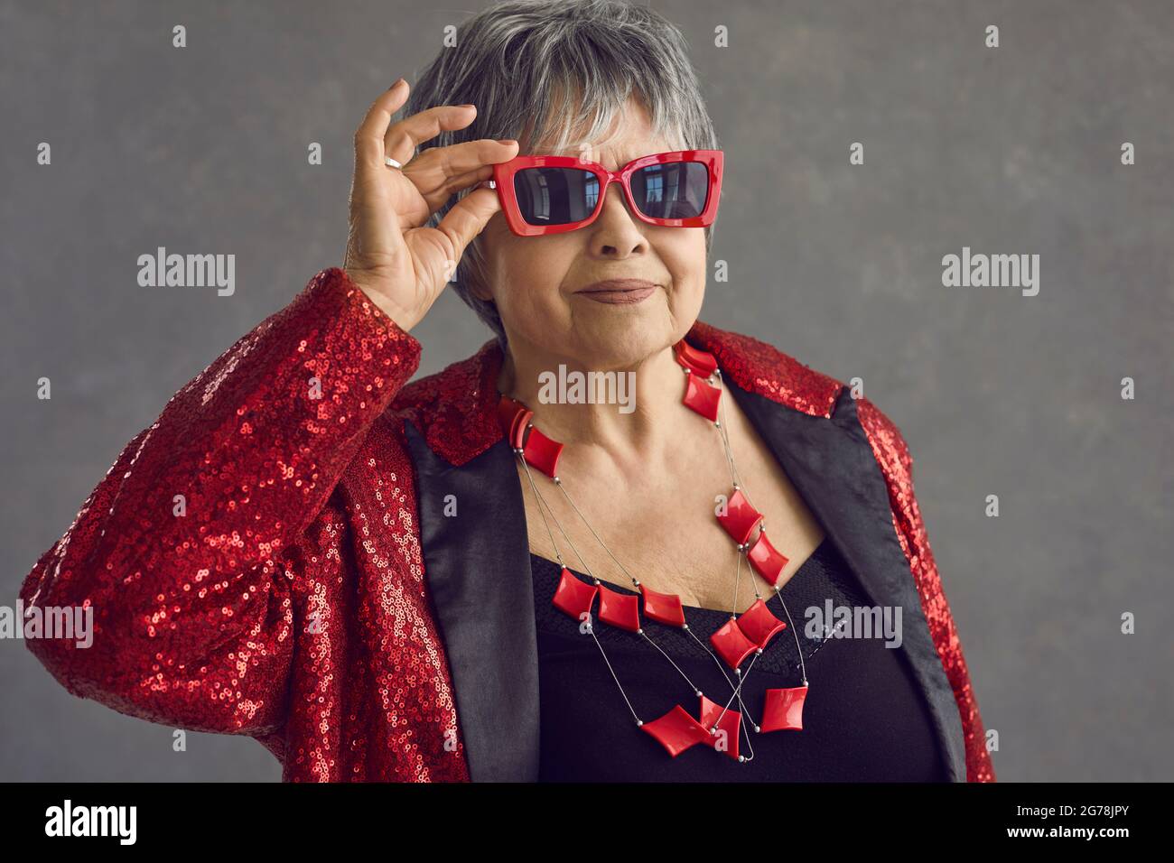 Porträt einer schönen älteren Frau in roter Sonnenbrille und Paillettenjacke, die im Studio posiert Stockfoto