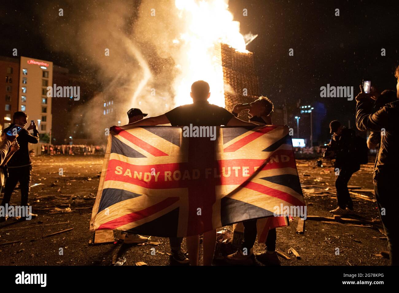 Ein Mann drapiert den Union Jack vor einem lodernden Scheiterhaufen am Lagerfeuer der Sandy Row in South Belfast um seine Schultern.am 11. Juli oder in der elften Nacht wird um Mitternacht im Norden Irlands ein Lagerfeuer angezündet, um die Wende vom 12. Juli zu feiern. In diesem Jahr jährt sich die Gründung Nordirlands zum hundertsten Mal und ist gleichzeitig der erste zugelassene Orangen-Marsch seit Beginn der Pandemie. Die Marschsaison ist in einer angespannten Zeit gefallen, in der die Brexit-Protokolle zu politischen Debatten führen und eine Spaltung der Gemeinschaft droht, die eine Rückkehr zu einer Volatilität droht, die seitdem nicht mehr zu beobachten ist Stockfoto