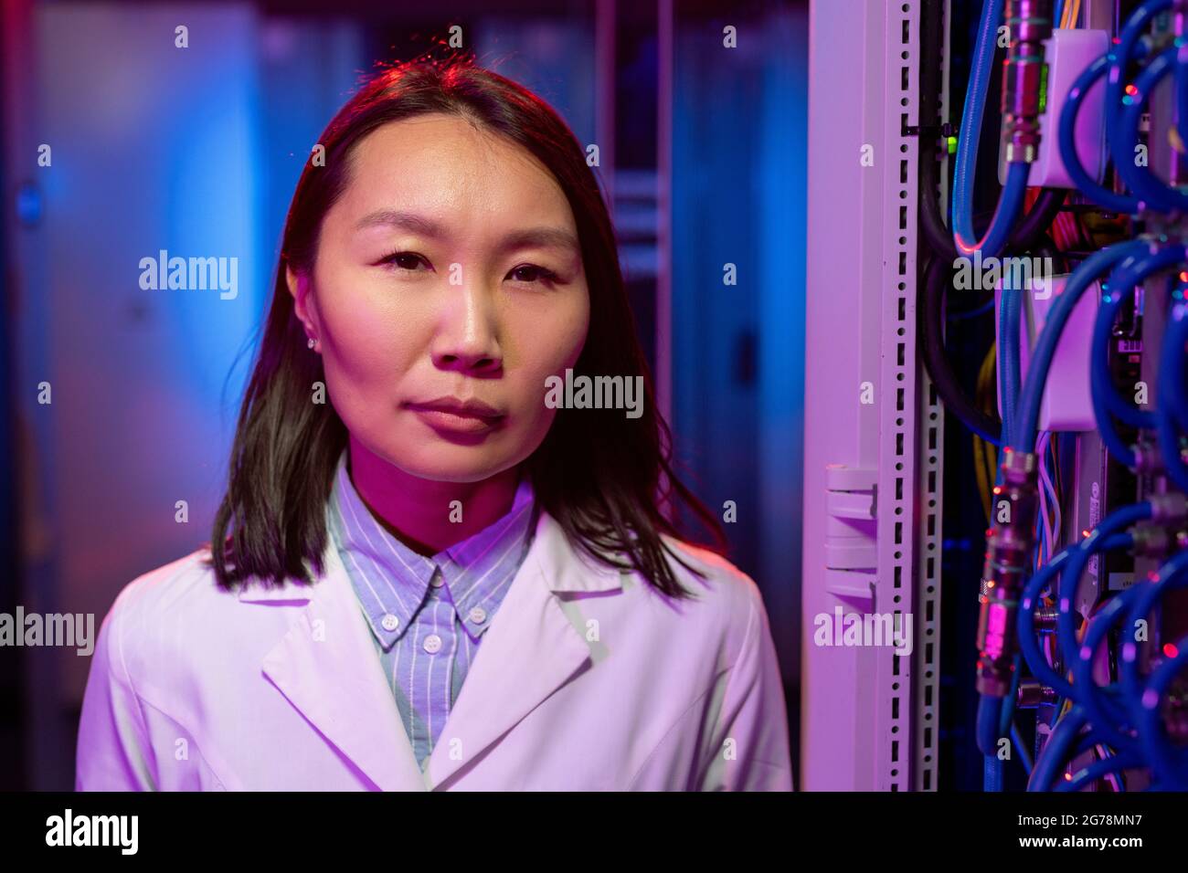Porträt eines selbstbewussten, qualifizierten, jungen asiatischen Serveringenieurs in weißem Mantel, der in der Nähe von Kabeln im Schrank steht Stockfoto