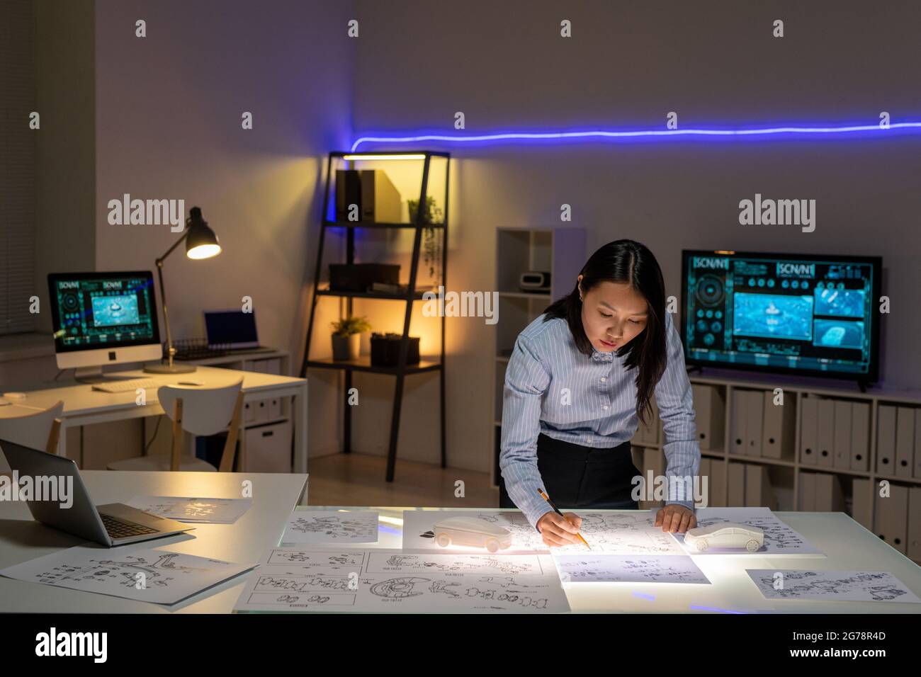 Konzentrierter junger asiatischer Autodesigner, der im dunklen Büro am Tisch steht und während der Deadline Skizzen zeichnet Stockfoto