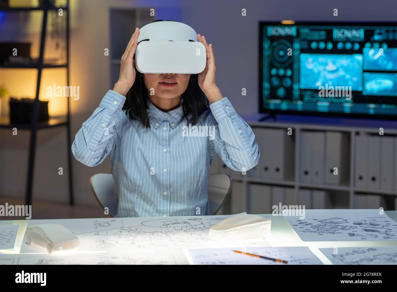 Junge asiatische Frau in blauem Hemd, die mit Skizzen am Schreibtisch sitzt und das Virtual-Reality-Headset anpasst, während sie sich auf die Arbeit vorbereitet Stockfoto