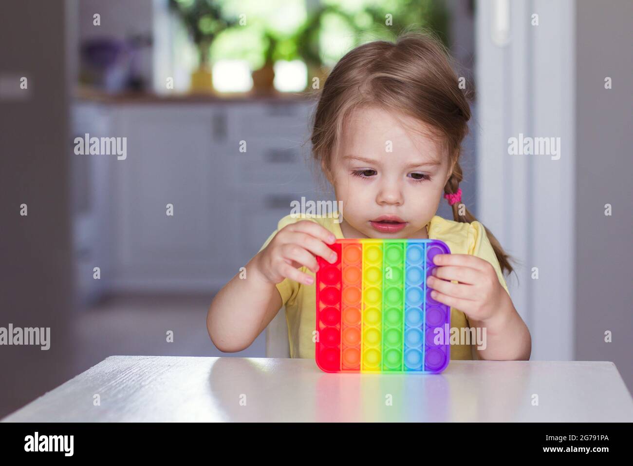 Kleine blonde Mädchen Kinder spielen mit neuen Trend sensorische Spielzeug Regenbogen Pop it. Antistress-Spielzeug für Kinder und Erwachsene.farbenfrohes Spielzeug einfache Grübchen. Verschmickt Stockfoto