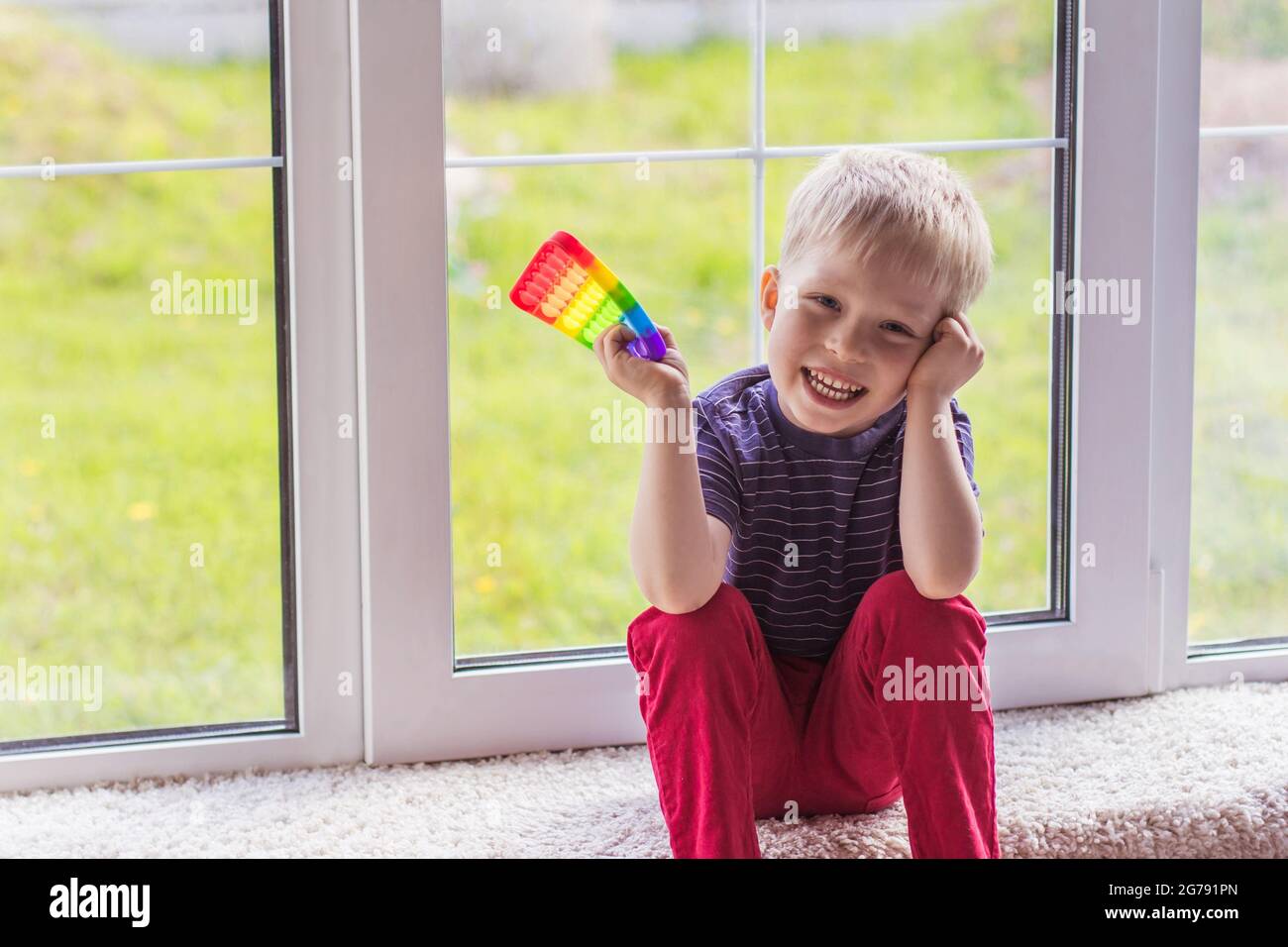Ein kleiner blonder Junge sitzt und schaut mit einem modernen bunten und hellen popit Spielzeug Simple Grübchen auf die Kamera. Das Kind mit bunten trendigen Antistress s Stockfoto