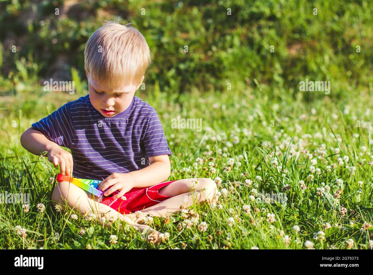 Kleiner Junge sitzt auf einem grünen Rasen und spielen Silikon-Spielzeug Antistress Pop it. Pop it Sinnesspielzeug. Stressabbau. Bunte Anti Stress Silikon sen Stockfoto
