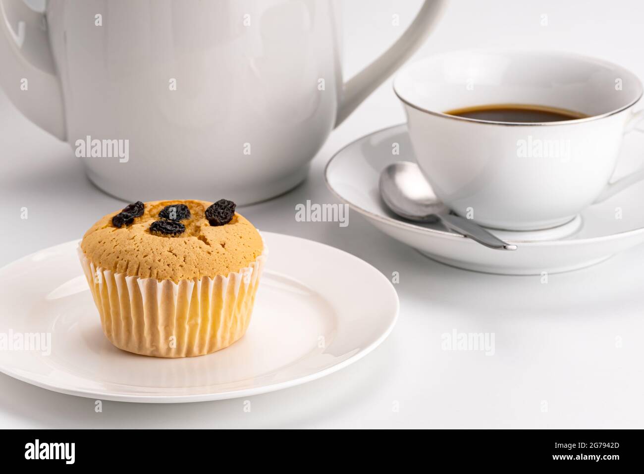Seitenansicht des einfachen Frühstücks mit Rosinenschwamm Cupcake in weißer Keramikschale und einer Tasse heißen schwarzen Kaffee mit Metalllöffel und einem Kaffeekocher auf w Stockfoto