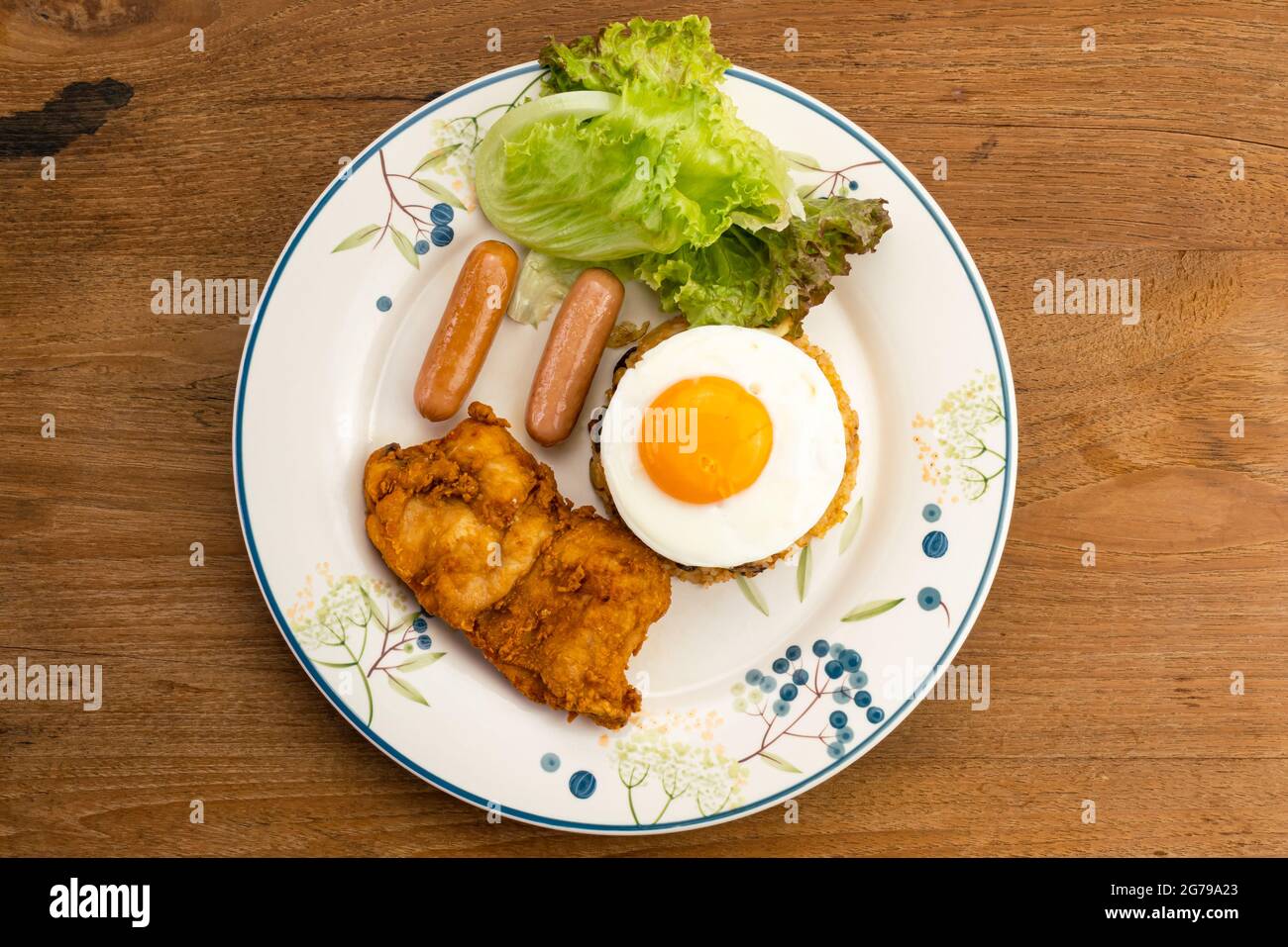 Draufsicht auf thailändischen amerikanischen frittierten Reis mit grünem Gemüse, Würstchen, Spiegelei, gebratenem Huhn und Rosinen in weißer Keramikschale auf Holztisch. Stockfoto
