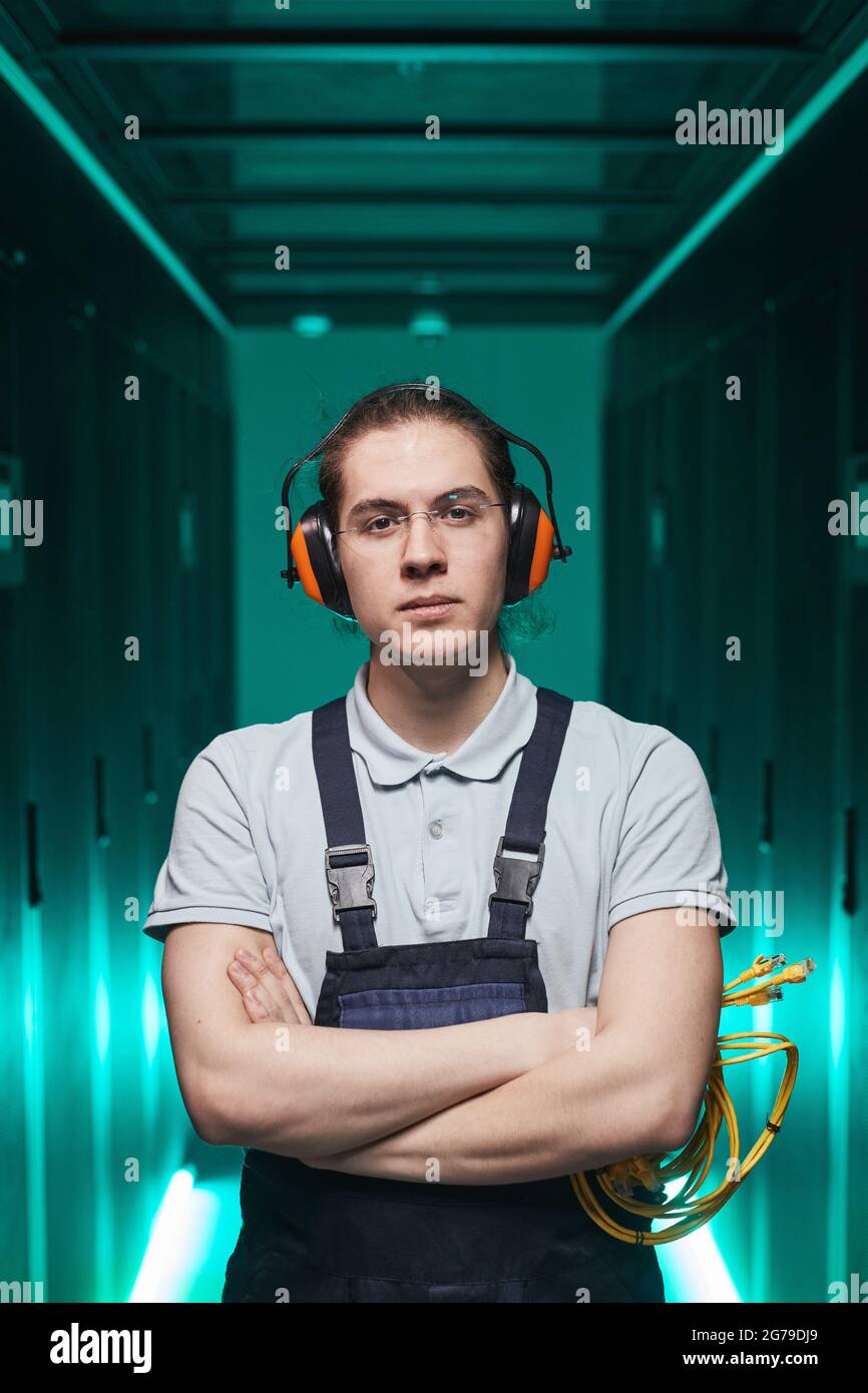Porträt eines jungen Netzwerktechnikers, der mit gekreuzten Armen im Serverraum auf die Kamera schaut Stockfoto
