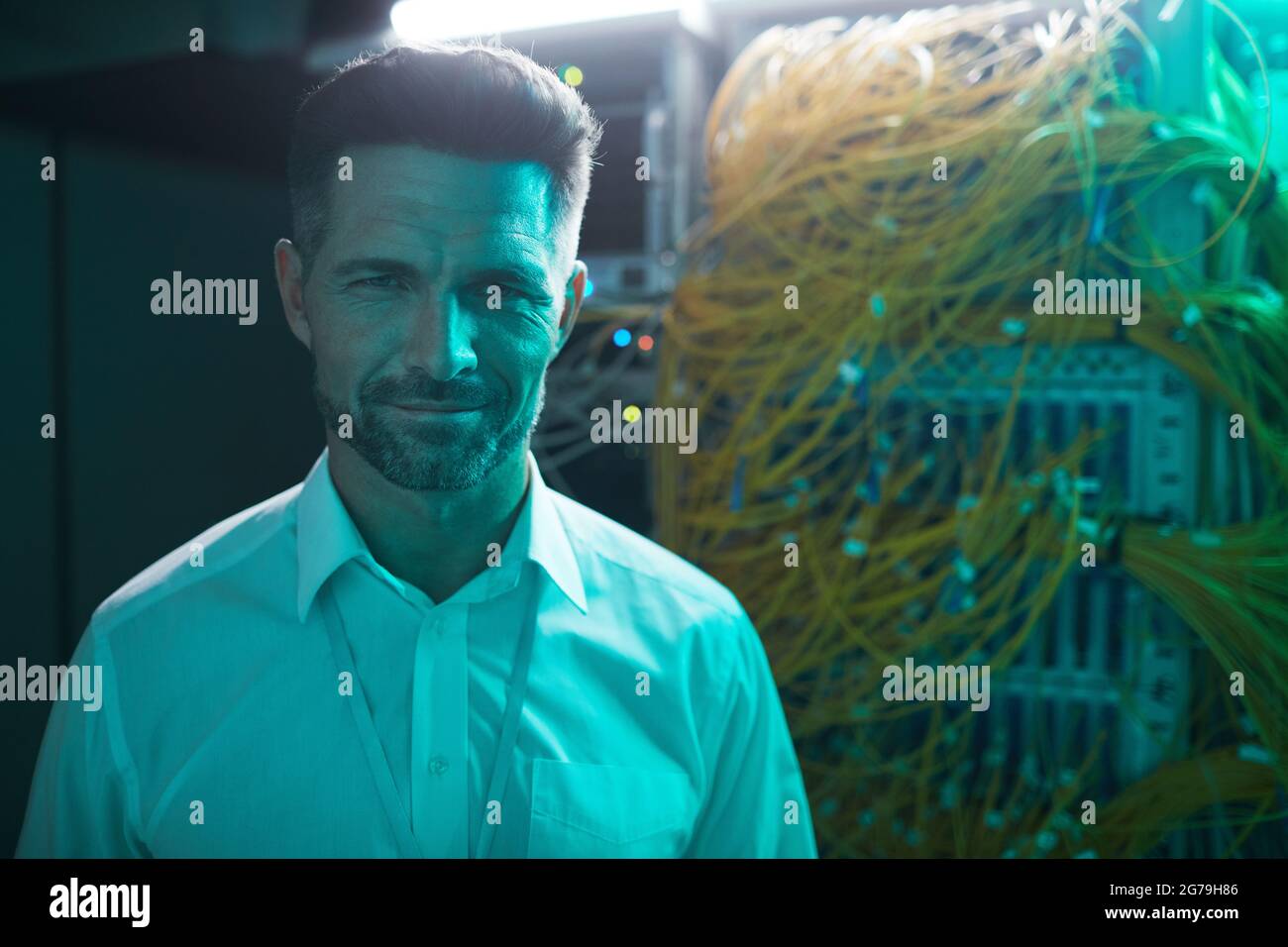 Porträt eines hübschen Dateningenieurs, der die Kamera im Serverraum anlächelt, während er mit einem Supercomputer in blauem Licht arbeitet und Platz für Kopien verwendet Stockfoto