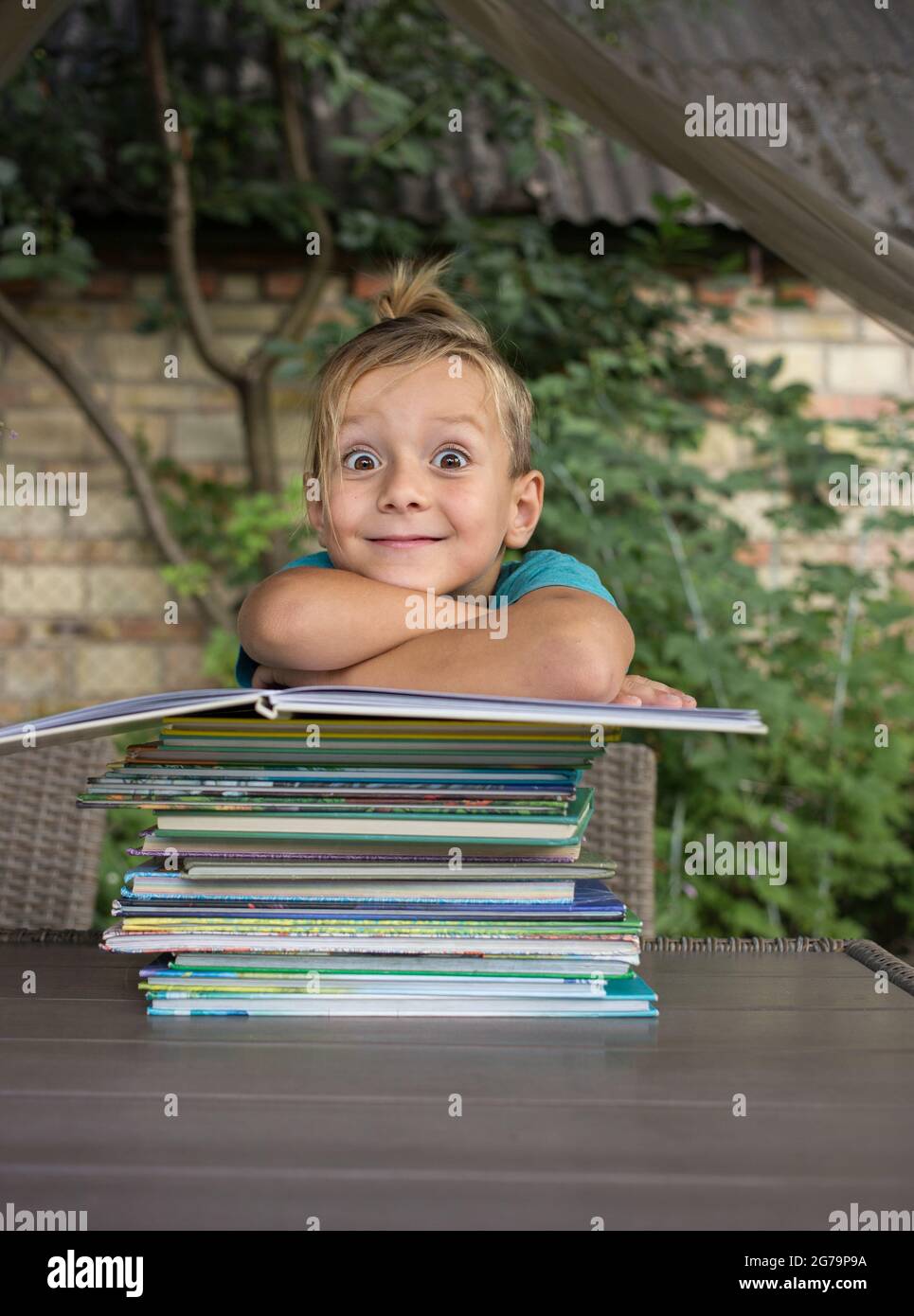 Witziger neugieriger Vorschulkinder sitzt an einem Tisch im Hof, vor ihm ist ein großer Haufen Kinderbücher. Bücher lesen, Kinderfantasien, ich Stockfoto