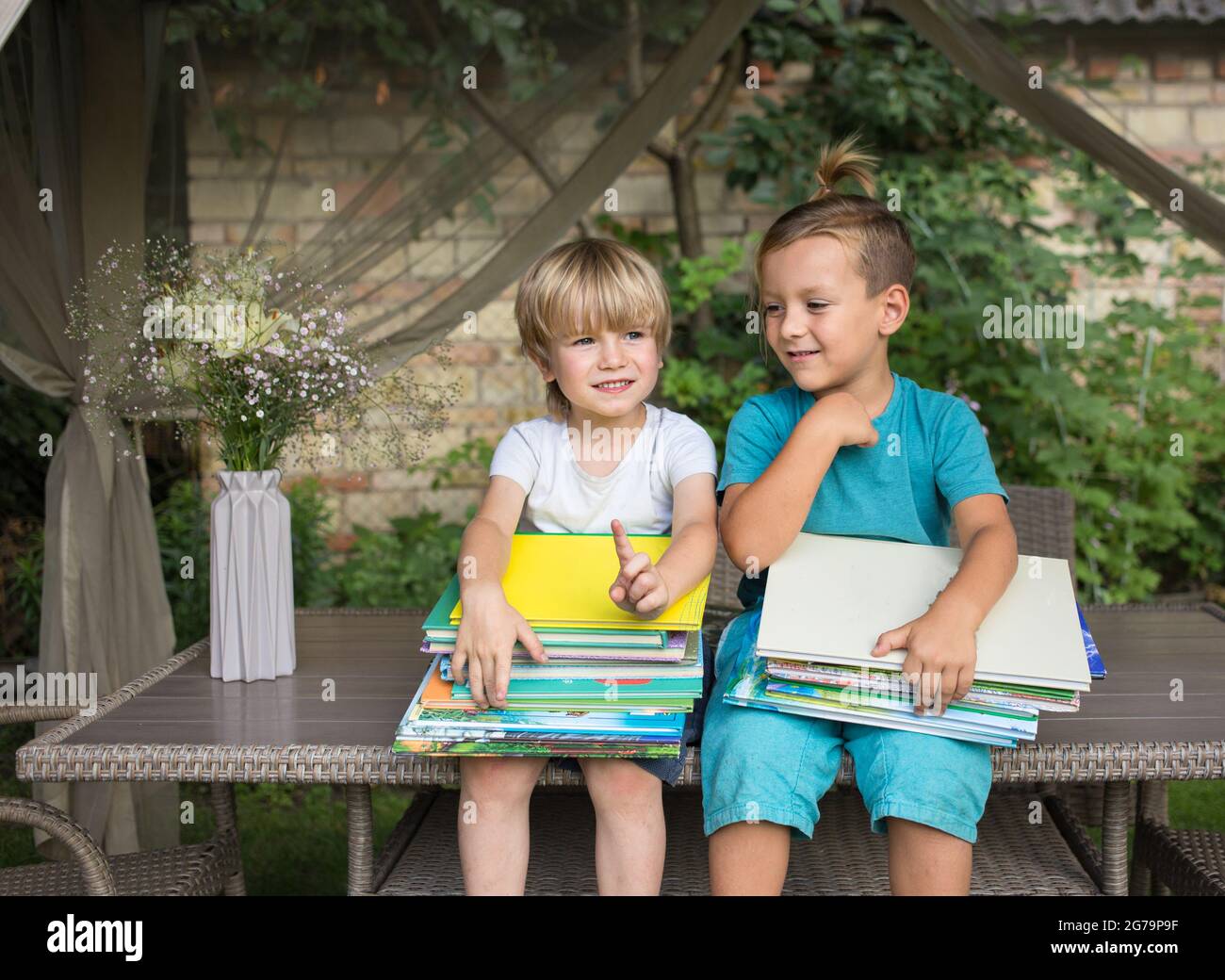 Zwei neugierige Vorschulkinder sitzen auf dem Tisch im Hof und halten Stapel von Kinderbüchern auf den Knien. Bücher lesen, Kinderfantasien, Kinderlichkeit, Stockfoto