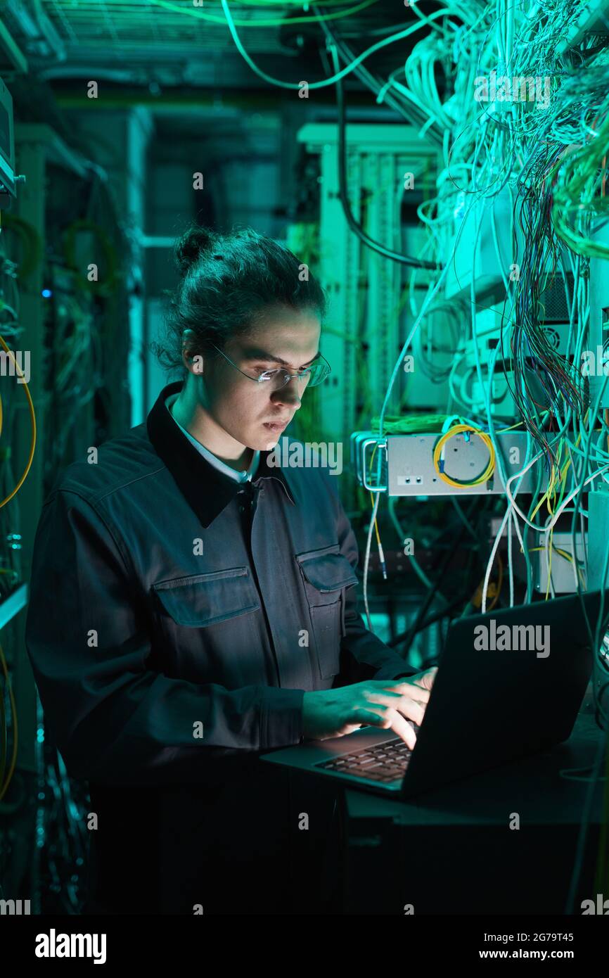 Vertikales Porträt eines jungen Netzwerktechnikers, der im Serverraum einen Laptop verwendet, während er im Rechenzentrum einen Supercomputer aufsetzt Stockfoto