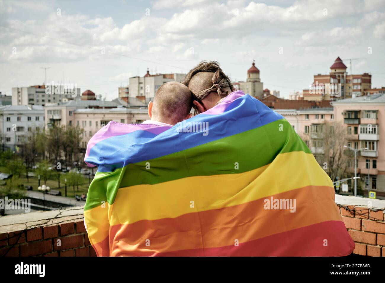 Rückansicht von zwei schwulen Männern, eingewickelt in Regenbogenfahne, die auf dem Dach stehen und die Stadt betrachten Stockfoto