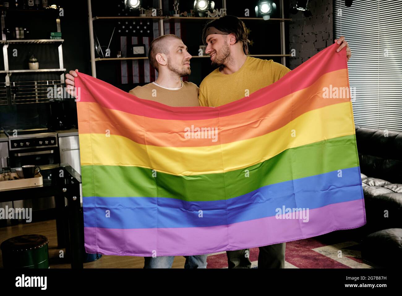 Junge liebevolle homosexuelle Männer halten Regenbogenfahne und schauen sich gegenseitig an, wenn sie in der Wohnung stehen Stockfoto