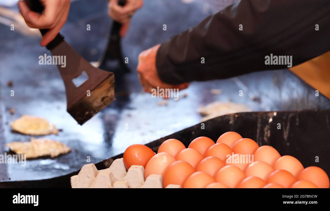 Ein Tablett oder Karton mit Eiern im Vordergrund mit einem Kochfeld bbq im Hintergrund. Hände und Kochutensilien drehen Spiegeleier. Spendenaktion für die Gemeinschaft Stockfoto