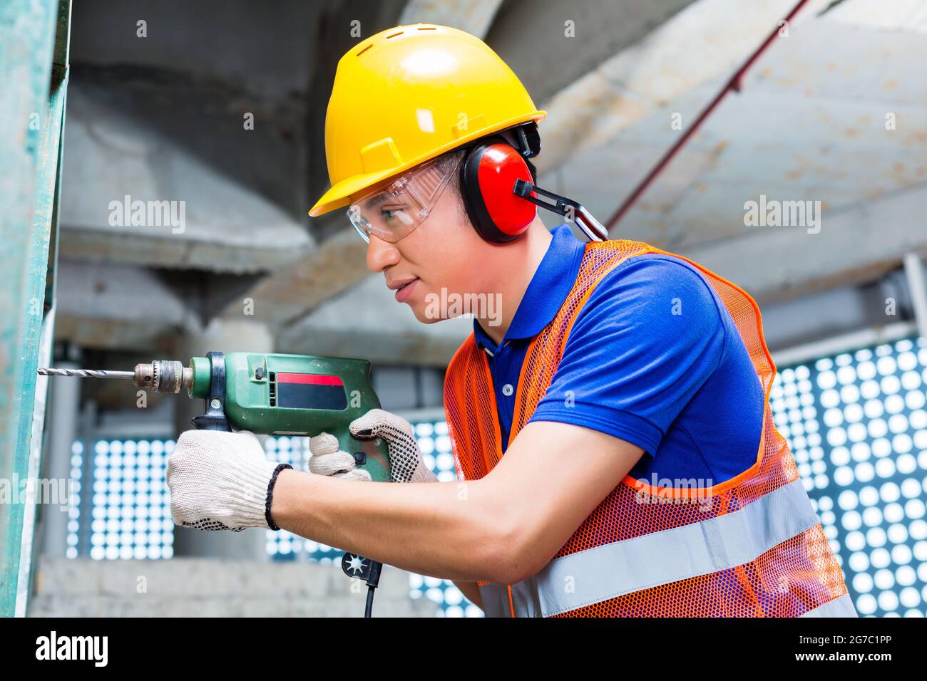Asiatisch-indonesischer Baumeister oder Arbeiter, der mit einer Maschine oder einem Bohrer, einem Gehörschutz und einem Hardhut oder Helm in einer Wand eines Turmgebäudes oder Konstruktis bohrt Stockfoto