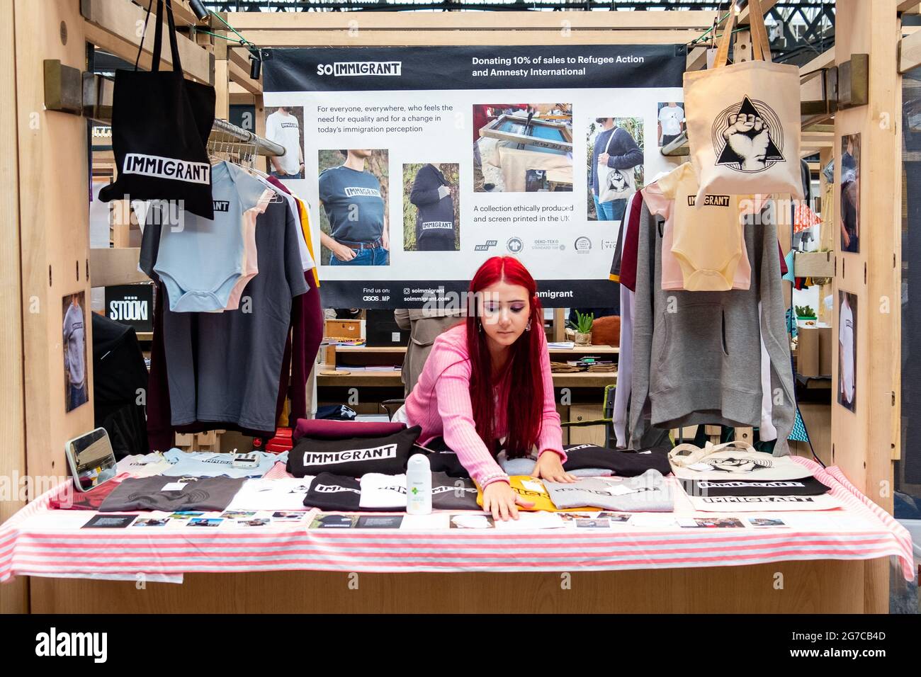 London - Juli 2021: Ein Mädchen, das eine Reihe von Modeartikeln auf dem Spitalfields-Markt in East London mit dem Wort / Branding ‘Immigrant’ verkauft Stockfoto