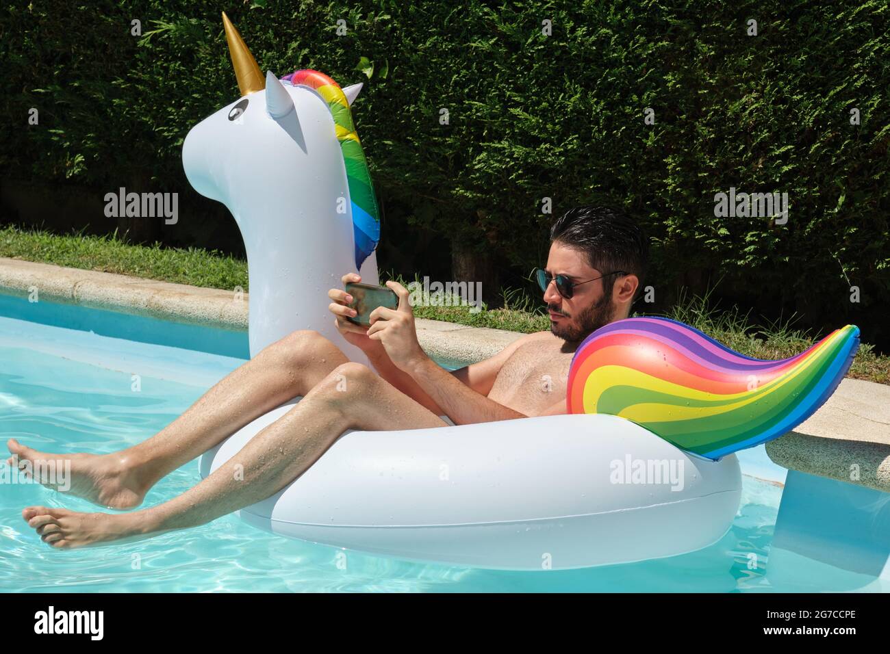 Junger Mann mit Sonnenbrille spielt am Telefon auf einem aufblasbaren Einhorn-Ring in einem Schwimmbad. Sommerkonzept. Stockfoto