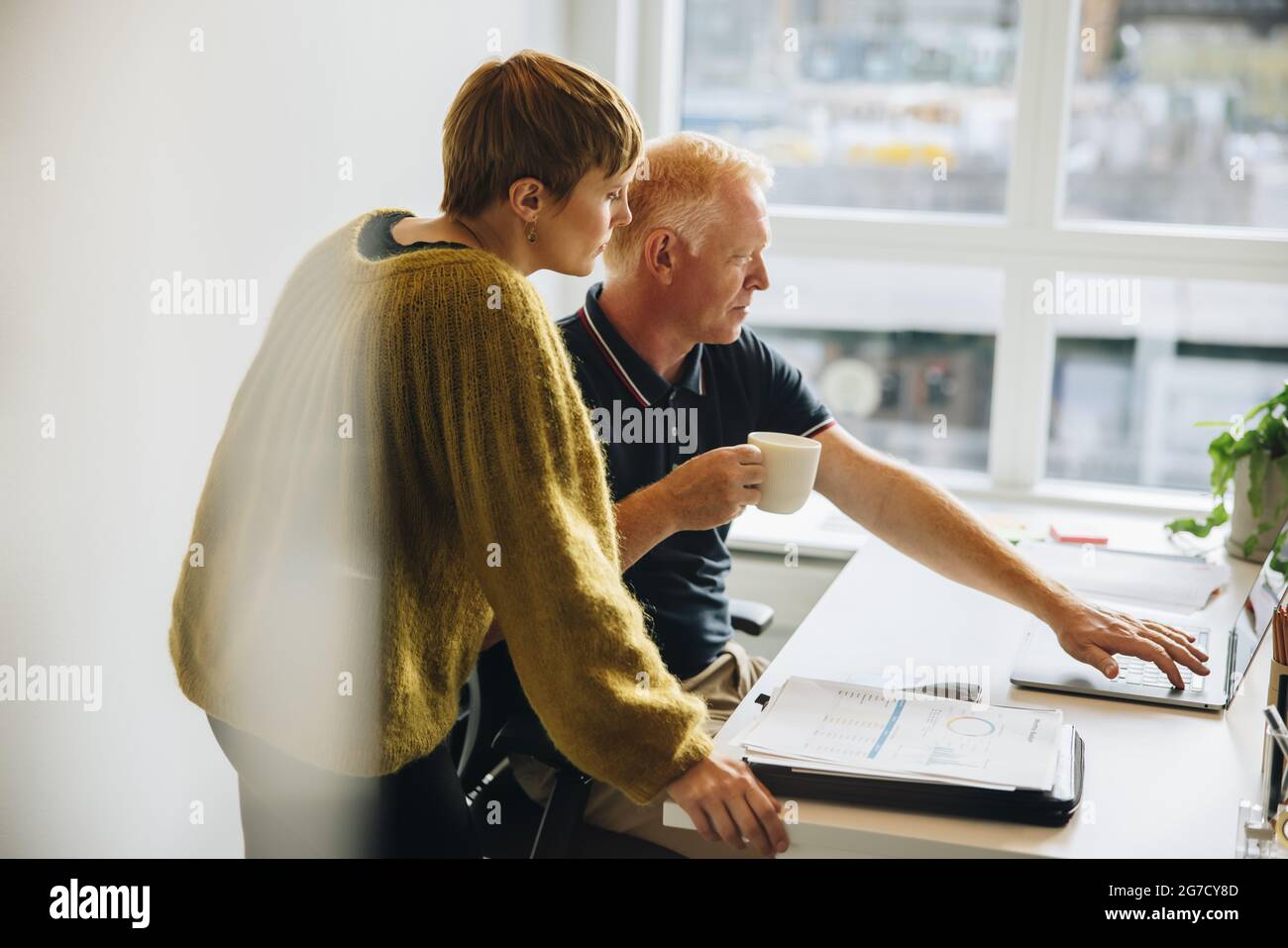 Zwei Geschäftsleute, die im Büro gemeinsam an einem Notebook arbeiten. Geschäftsmann hält Kaffee und zeigt seinem Geschäftspartner etwas auf dem Laptop. Stockfoto