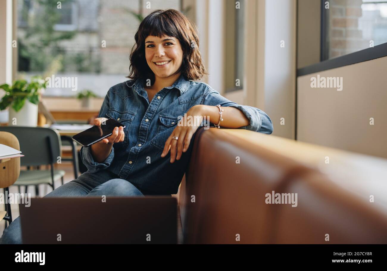 Geschäftsfrau, die auf dem Sofa sitzt und im Büro in die Kamera schaut. Freiberuflicher Mitarbeiter, der im Coworking Space arbeitet. Stockfoto