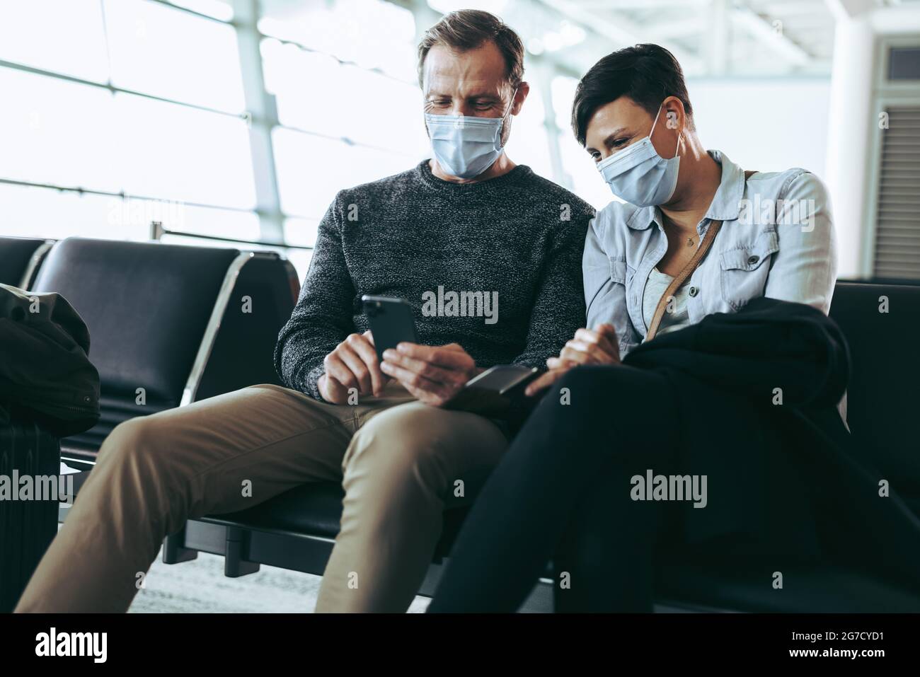 Mann zeigt der Frau am Flughafen sein Handy. Setzen Sie sich mit dem Telefon in Gesichtsmasken, während Sie auf ihren Flug warten. Stockfoto