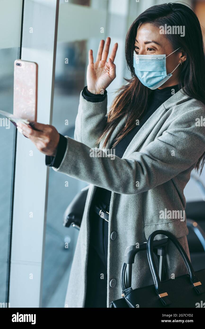 Frau trägt Gesichtsmaske Videoanruf mit ihrem Handy. Weibliche Reisende, die einen Videoanruf in der Wartelounge des Flughafens führen. Stockfoto