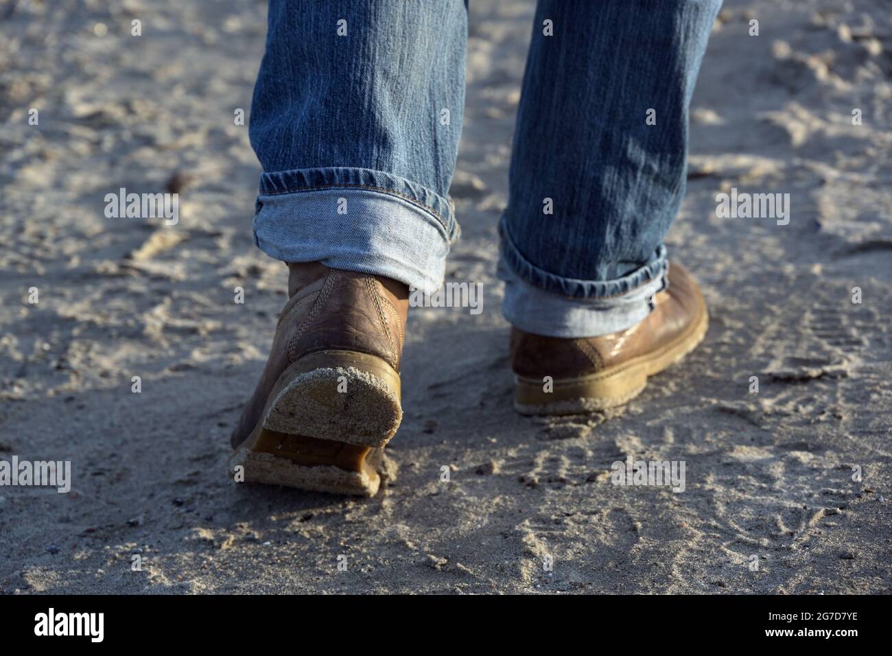 Füße eines Mannes mit Lederstiefeln und aufgerollten Jeans, die am Strand auf dem Sand vorwärts laufen, Freizeitangebot, ausgewählter Fokus, enge Tiefe von f Stockfoto