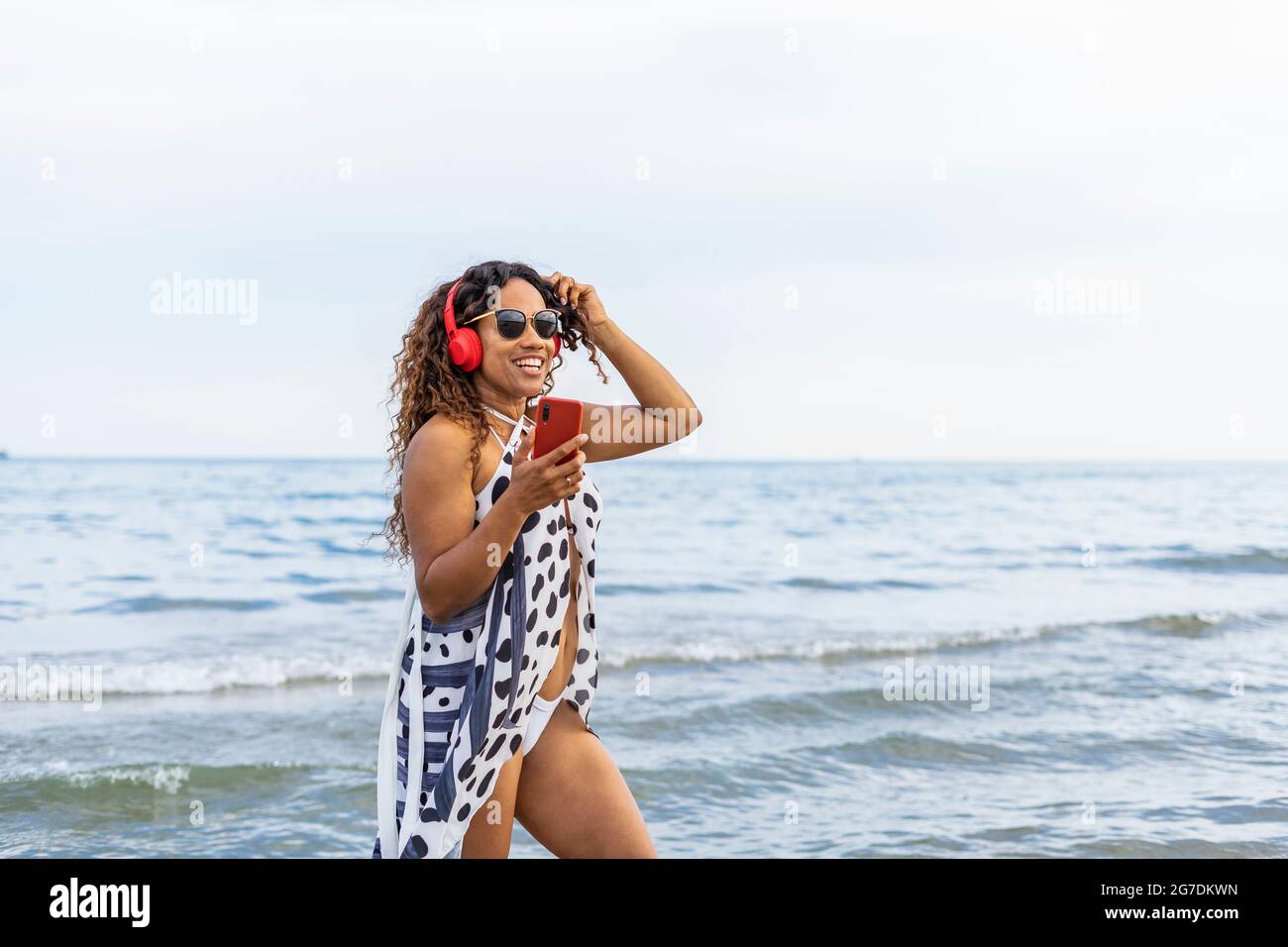 Portrait lächelnde afroamerikanische Frau, die am Strand bei Sonnenuntergang Musik hört und tanzt.Frau, die rotes Smartphone benutzt und den Sommertag am Strand genießt Stockfoto