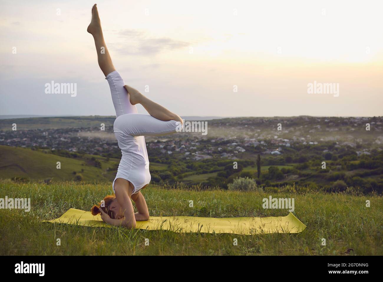 Frau, die bei Sonnenuntergang im grünen Sommerfeld Yoga praktiziert, während sie Kopfstand-Übungen macht Stockfoto