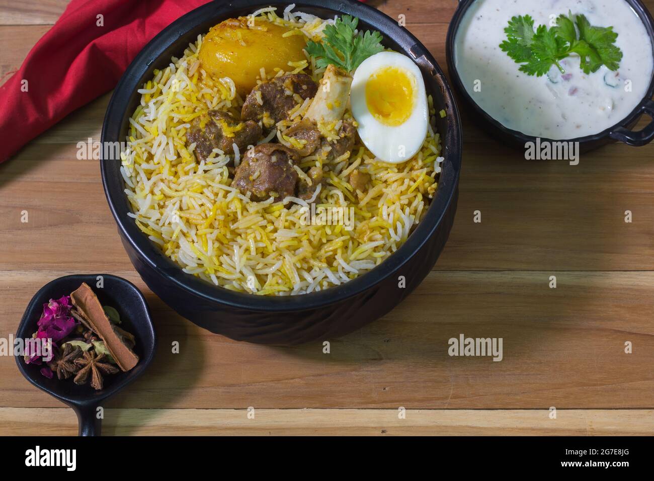 Köstliche Hammelfleisch Biryani serviert in schwarzem Topf mit Raita. kalkutta Stil Dum Biryani mit Hammelfleisch, gekochtem Ei und Kartoffeln. Holzhintergrund. Traditionelle m Stockfoto