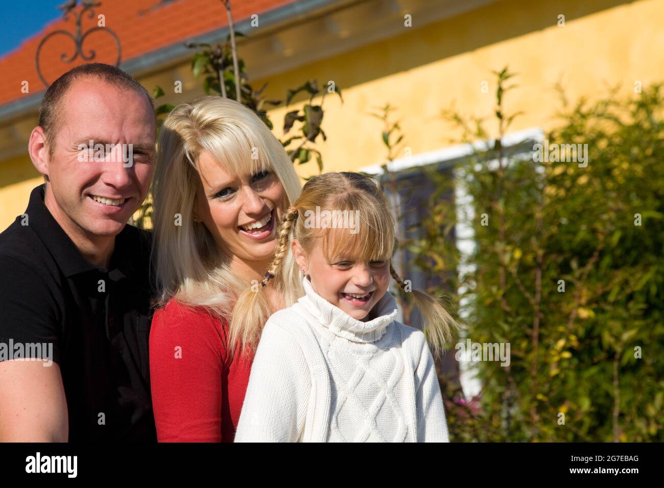 Ein junges Paar mit ihrer Tochter sitzt an einem perfekten Tag vor ihrem gelben Haus Stockfoto
