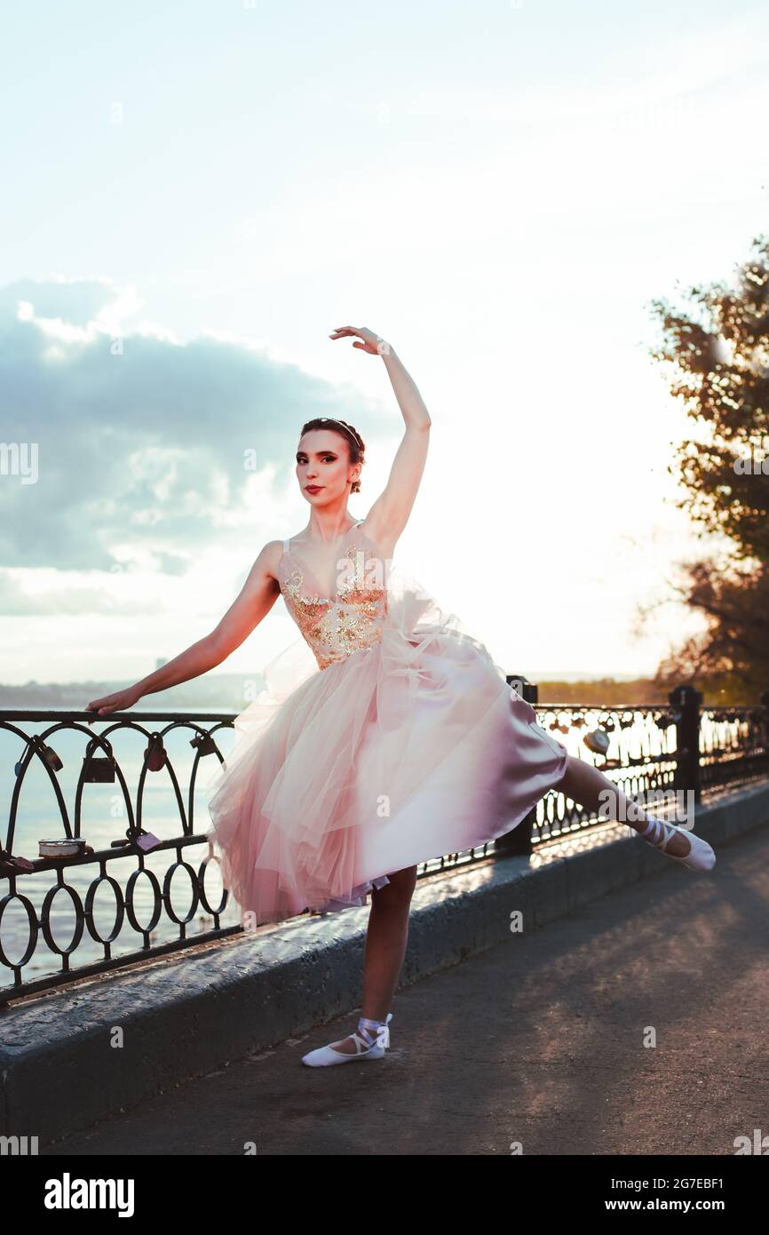 Eine dünne Ballerina in einem pinken Seiden-Korsett-Kleid hebt ihr bein mit spitzenzägen anmutig an den Flussufer in einem gusseisernen Zaun in der Stadt zurück Stockfoto