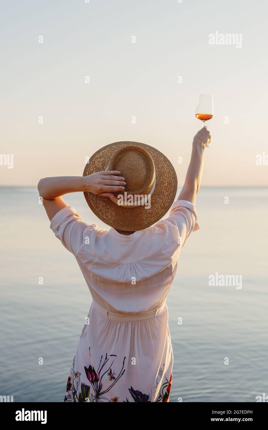 Rückansicht einer schönen Frau im Strohhut, die ein Glas Wein mit Meer im Hintergrund genießt. Stockfoto