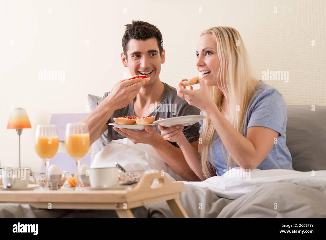 Junges Paar feiern mit Frühstück im Bett sitzen Essen berry Jam o Toast, begleitet von frischen Orangensaft und gekochte Eier. Stockfoto