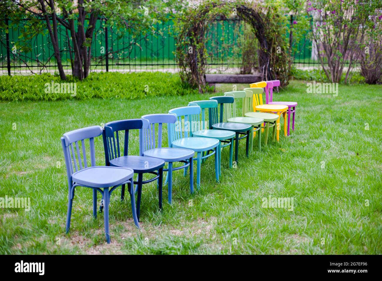 Eine diagonale Reihe von Regenbogen-Holzstühlen auf einer grünen Wiese im Park, die sich auf eine Familienfeier an einem Wochenende im Freien vorbereiten Stockfoto