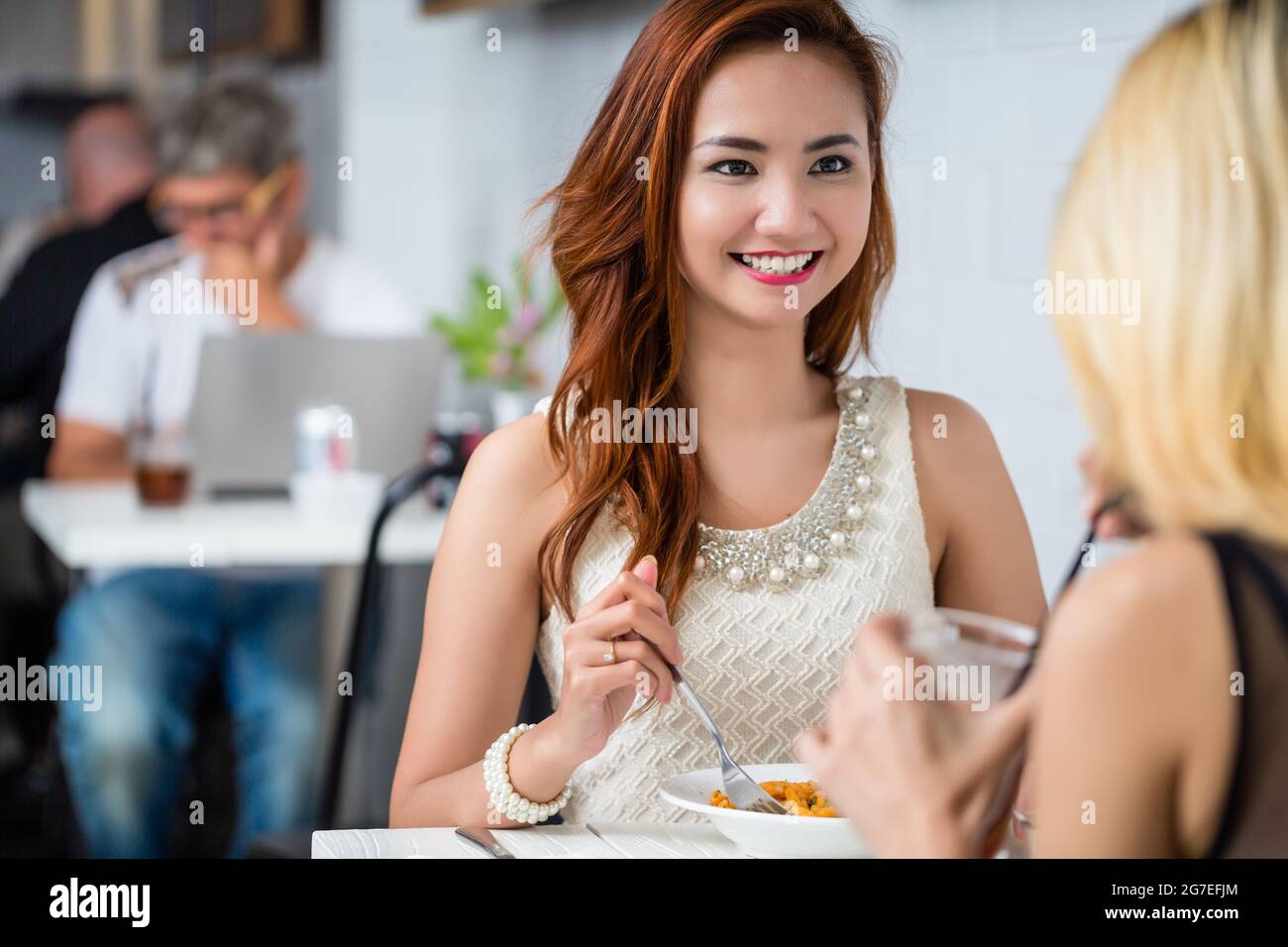 Attraktive, elegante junge asiatische Frau, die mit einem Freund in einem Restaurant zusammen lächelnd und über der Schulter am Tisch sitzt und sich unterhält Stockfoto