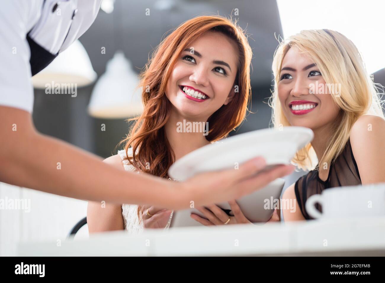 Zwei lächelnde, attraktive asiatische Frau, die in einem Restaurant mit einem Teller Essen serviert wird, aus dem Winkel an den Kellnern vorbei, mit Fokus auf ihre Gesichter Stockfoto